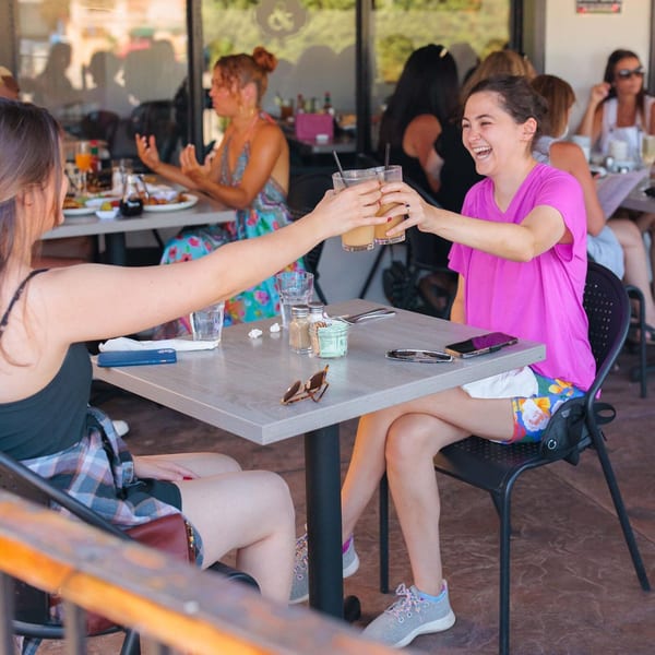 Two women clinking glasses of juice at an outdoor table, with other diners in the background.
