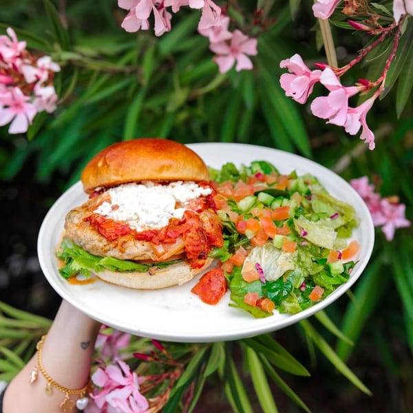 A hand holds a plate with a chicken burger topped with feta and tomato sauce, and a side salad.