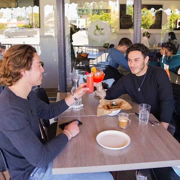 Two men at Leo & Lily restaurant clinking glasses with colorful drinks, food, and coffee on the table.