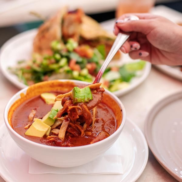 A hand spoons red tortilla soup with avocado and tortilla strips from a white bowl at Leo & Lily.