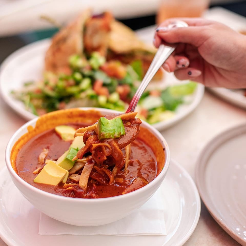 A hand spoons red tortilla soup with avocado and tortilla strips from a white bowl at Leo & Lily.