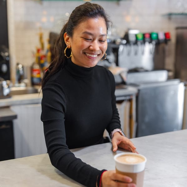 Smiling Leo & Lily barista in a black turtleneck serving a latte in a to-go cup.