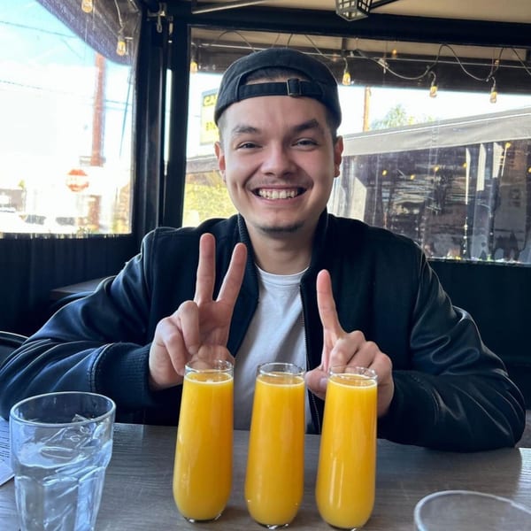 A smiling man in a black cap and jacket makes peace signs behind three mimosas at Leo & Lily.