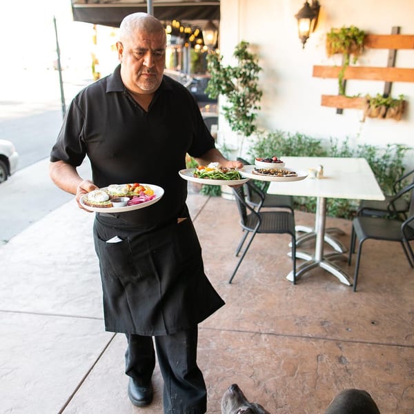 A server in a black uniform carries two plates of breakfast food on an outdoor patio.