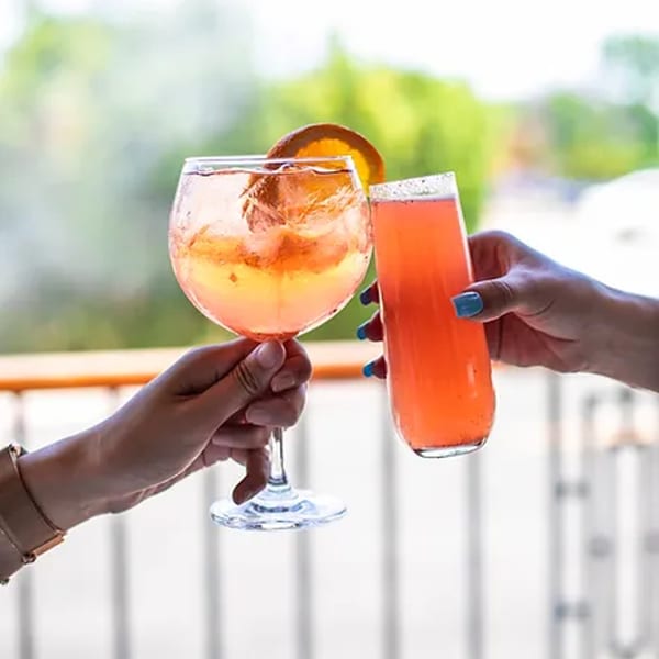 Two hands clinking glasses of orange and pink cocktails with a blurred green background.