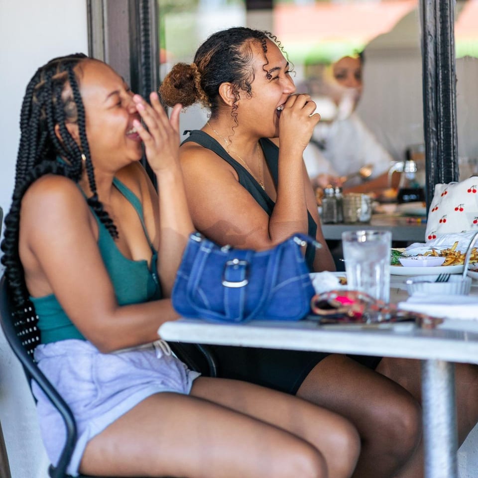 Two women laughing joyfully while dining outdoors at Leo & Lily, with breakfast dishes on the table.
