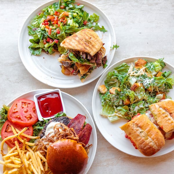 Three plates of Leo & Lily's breakfast items: a panini with salad, a burger with fries, and a sandwich with Caesar salad.