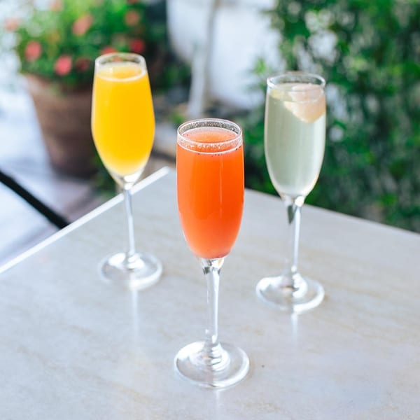 Three colorful mimosas, orange, red, and light green, in champagne flutes on a white table.